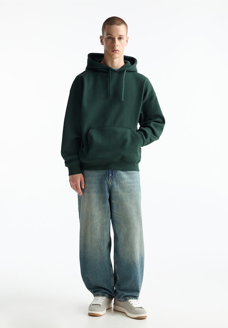 Grüner Kapuzenpullover mit einer Vordertasche, kombiniert mit lockeren hellblauen Jeans und grauen Sneakern, vor einem weißen Hintergrund.