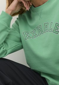 Sudadera verde con la palabra "EXHALE" en un texto curvado y en negro contrastante. El tejido tiene una textura suave con puños de canalé.