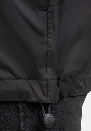 Veste extérieure noire en tissu lisse et texturé. Cordon de serrage à l'ourlet avec un bouton rond. Superposée à un vêtement sombre à motifs.