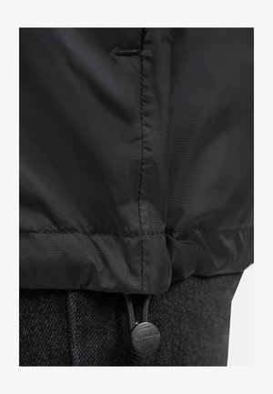 Veste extérieure noire en tissu lisse et texturé. Cordon de serrage à l'ourlet avec un bouton rond. Superposée à un vêtement sombre à motifs.