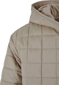 Beige quiltet pufferjakke med høj krave og blødt, tekstureret interiør. Distinkt gittermønster med synlige stingdetaljer.