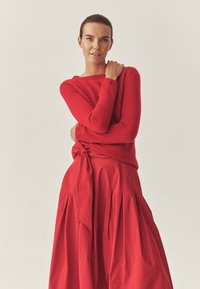 Sarkans adīts džemperis, apvienots ar plūstošu sarkanu svārku. Džemperim ir garas piedurknes un apaļa kakla izgriezums. Svārkiem ir krokas un saistīta jostas detaļa.