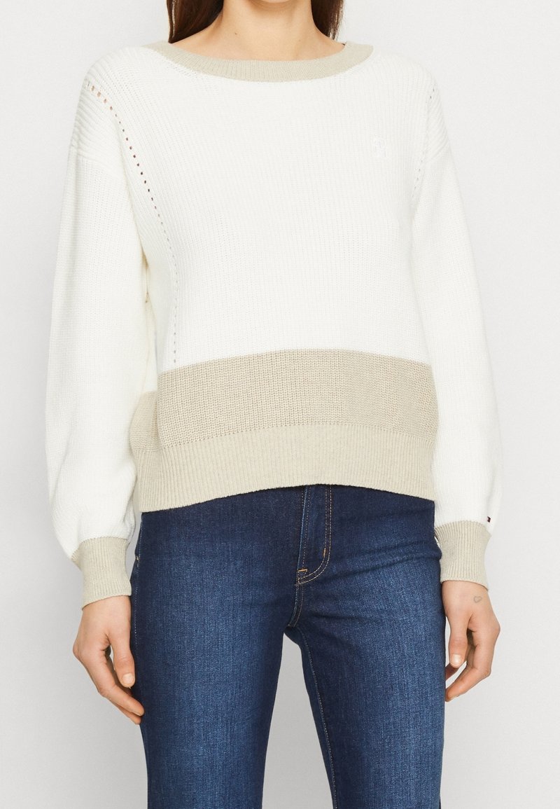 Femme portant un pull en maille blanc avec des poignets et un ourlet beige, associé à un jean bleu foncé, se tenant devant un fond uni.