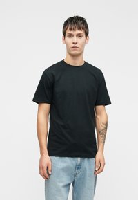 Czarna bawełniana koszulka z krótkim rękawem, okrągłym dekoltem i luźnym krojem. Model nosi jasnoniebieskie dżinsy. Prosty design, bez wzorów.