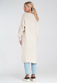 Kobieta z blond włosami zwi�ązanymi w kucyk, ubrana w długi beżowy sweter, jasnoniebieskie jeansy i trampki w panterkę, stojąca bokiem na jednolitym tle.