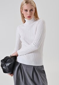 Donna con capelli biondi indossa un maglione a collo alto bianco strutturato e pantaloni grigi plissettati, tiene una borsa in pelle nera contro uno sfondo grigio.
