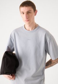 Lys blå bomulds T-shirt med korte ærmer, der har et diskret "HUGO" logo. Modellen holder en sort stofpose under den ene arm.
