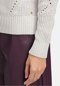 Helle beige gestrickte Pullover mit gerippten Bündchen, ausgestattet mit einem kleinen goldenen Logo und einem strukturierten Muster. Über einem dunkelvioletten Kleidungsstück getragen.