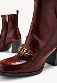 Bottes courtes bordeaux brillantes avec un accent de chaîne dorée, dotées d'un bout carré et d'un talon carré. Texture lisse et détail de fermeture éclair latérale.