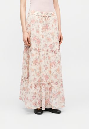 Femme portant une longue jupe fluide crème avec imprimé floral rose, volants à étages, taille à cordon de serrage, et chaussures plates noires.