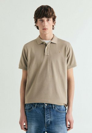 Polo beige con cuello clásico, mangas cortas, un logo sutil en el pecho y una textura de tela suave, combinado con vaqueros azules.