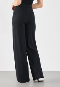 Mujer con pantalones negros acampanados de tiro alto y costura de canalé, usando zapatos blancos, de pie con una mano en la cadera frente a un fondo liso.