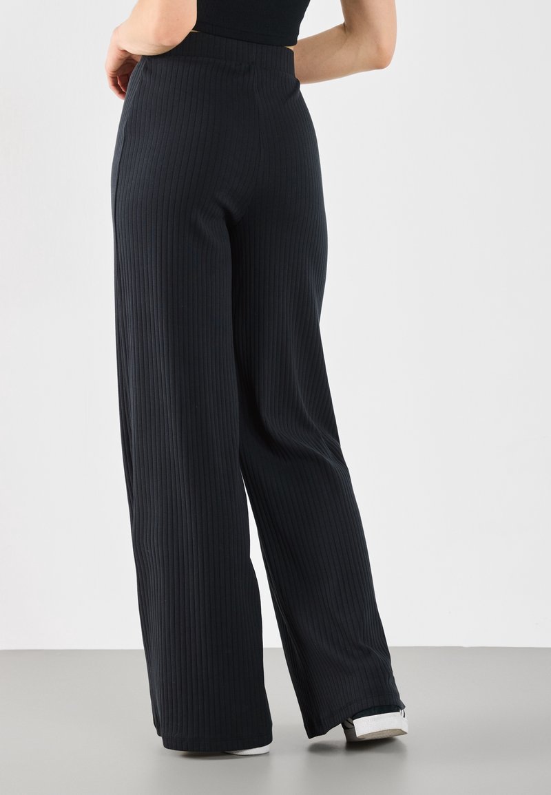 Mujer con pantalones negros acampanados de tiro alto y costura de canalé, usando zapatos blancos, de pie con una mano en la cadera frente a un fondo liso.