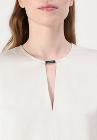 Blouse beige clair avec une texture lisse, présentant un col en forme de trou et une petite étiquette noire près du col. Design minimaliste.