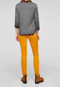 Person, der senfgelbe Hosen, braune Schnürstiefel und einen grauen Pullover über einem hochgerollten orangefarbenen Karoshirt trägt, steht mit dem Rücken zur Kamera.