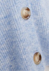 Ljusblått stickat tyg med två stora genomskinliga beige knappar sydda på, som visar detaljerad textur och sömnadsmönster.