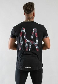 Svart t-shirt med en stor grafik av ett 'A' fyllt med skallar och rosor, korta ärmar med mönstrade detaljer, och en nedre etikett som säger "LIMITÉ."