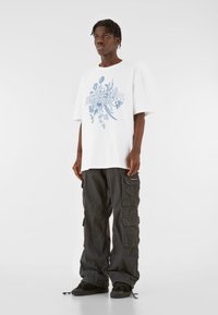 Hvid oversized t-shirt med blåt blomstermotiv, kombineret med løstsiddende sorte cargo-bukser med flere lommer og stropper.