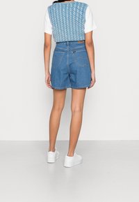 Sininen neuleliivi valkoisen t-paidan päällä, yhdistetty korkeavyötäröisiin sinisiin farkkushortseihin ja valkoisiin lenkkareihin. Shortseissa on rento istuvuus ja taskut.