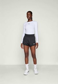 Bjela dugih rukava sportska majica s Adidas logotipom, uparena s crnim kratkim hlačama s elastičnom gumom u struku i bijelim Adidas akcentima, nosi se uz bijele tenisice.