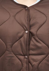 Κοντινή λήψη καφέ υφάσματος με καπιτονέ σχέδιο, με τρία κουμπιά κούμπωμα και εμφανείς ραφές κατά μήκος των άκρων και των ραφών.