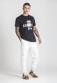 Černé tričko s kresleným vzorem, bílé cargo kalhoty s kapsami a hnědé tenisky s leopardím vzorem. Jednoduché pozadí.