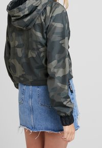 Chaqueta con capucha recortada en patrón de camuflaje verde con puños elásticos, combinada con una falda de mezclilla desgastada. Presenta detalles de cremallera.