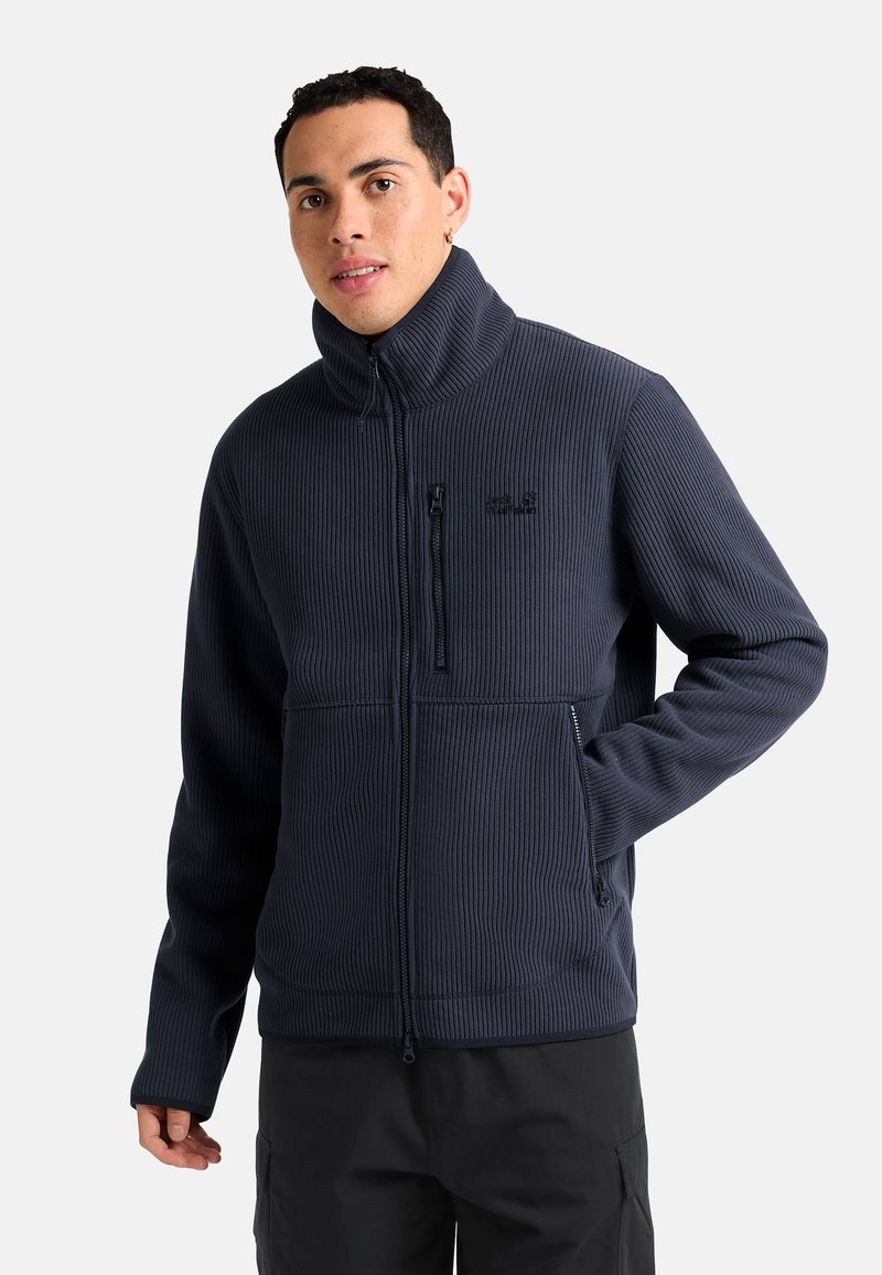 Giacca in pile a coste blu navy con colletto alto, tasche con zip e dettagli del logo neri. Caratterizzata da una vestibilità aderente e un tessuto testurizzato.