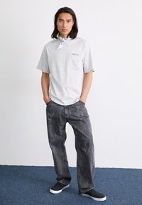 Grå bomull t-skjorte med korte ermer, med en liten logo på brystet. Svarte mønstrete jeans med mørke detaljer og svarte slip-on sko.