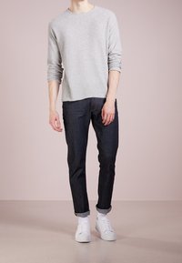 Hellgrauer, gestrickter Pullover mit langen Ärmeln, kombiniert mit dunklen Jeans und weißen Sneakers. Die Jeans haben hochgerollte Säume.