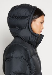 Moteris profiliu, vilkinti juodą pūkinę striukę su kapišonu, užtrauktą iki smakro, prieš paprastą šviesią fono.