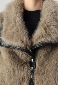 Faux fur jas met een luxe beige textuur, voorzien van zwarte leren accenten en een sjawl kraag, afgesloten met zilveren knopen.