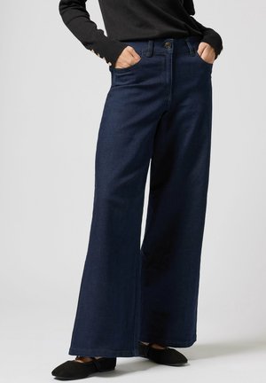 Person iført mørkeblå viddebukser, sort langærmet top med knaplukning ved manchetter og sorte flade sko, hænder i lommerne.