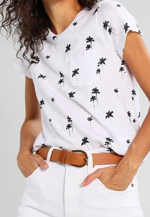 Nainen, jolla on päällään valkoinen t-paita, jossa on mustia palmupuun kuvia, sisällytetty valkoisiin housuihin, joissa on ruskea nahkavyö, kädet taskuissa.