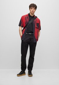 Short-sleeve shirt in red and black with abstract pattern, paired with black pants and black sneakers featuring gum soles. Adjustable strap across the chest.