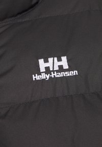Černá prošívaná bunda z materiálu s bílým vyšitým logem "HH" a "Holly-Hansen" na přední straně. Hladká textura, minimalistický design.