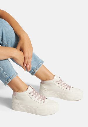 Persona que lleva zapatillas de caña alta de color rosa claro con suelas gruesas y cordones rosas, combinadas con vaqueros azul claro remangados.