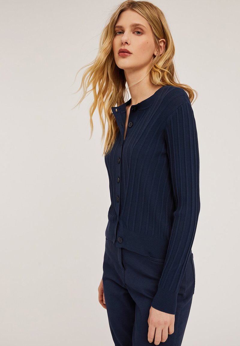 Granatowy cardigan w prążki z długimi rękawami, okrągłym dekoltem oraz zapięciem na guziki z przodu. Gładka tekstura i dopasowany fason.
