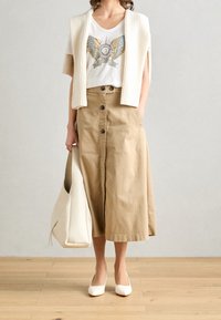 Béžová midi sukně na knoflíky je zkombinována s bílým tričkem s potiskem a krémovým žebrovaným kardiganem. Doplňky zahrnují bílou tašku a lodičky.