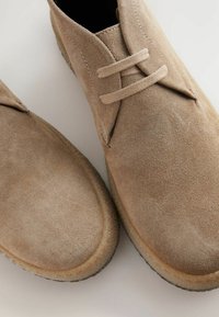 Bottines en suède beige avec fermeture à lacets, bout arrondi et surface texturée. Semelle en caoutchouc avec un léger surpiqûre contrastée.