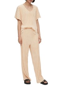 Tenue plissée de couleur pêche comprenant un top à col en V et manches courtes, assortie à un pantalon droit, le tout associé à des sandales à glissière noires.