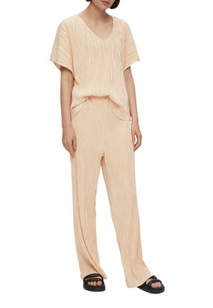 Perzikkleurige, geplooide outfit met een V-hals top met korte mouwen en rechte pijpen, gecombineerd met zwarte instapsandalen.