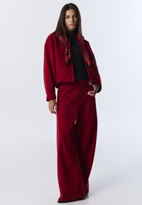 Chaqueta roja corta con un cuello de tortuga negro, combinada con pantalones anchos rojos. Presenta una bufanda con un patrón rojo y negro en el cuello.