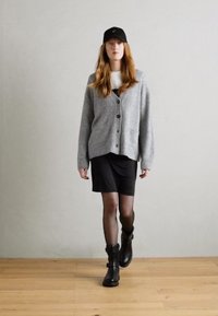 Cardigan gris texturé avec des boutons, robe noire, collants noirs transparents et bottines noires, associés à une casquette noire. Cadre intérieur.