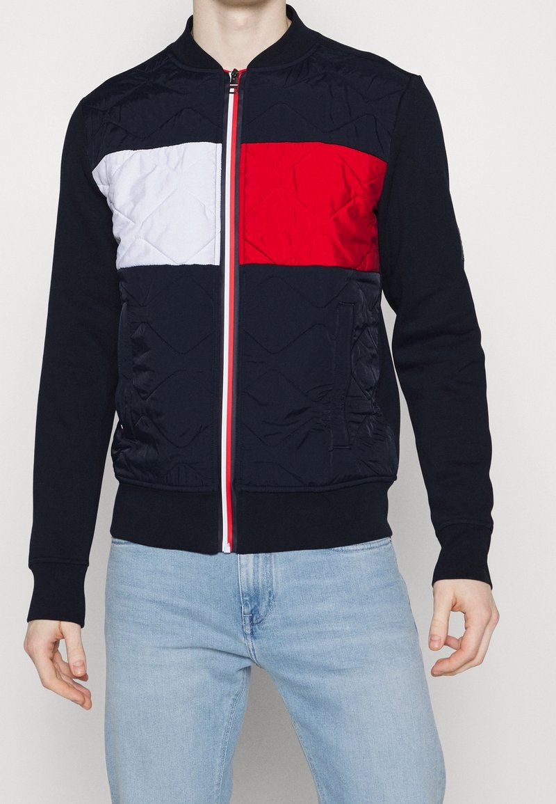 Veste zippée pour homme avec un design en color-block présentant des panneaux bleu marine, rouge et blanc, une texture matelassée et un détail de fermeture éclair contrastant.