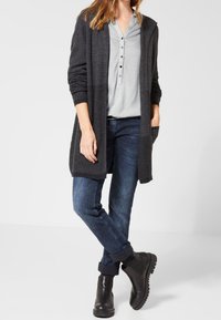 Cardigan gris en maille à manches longues, associé à un chemisier léger à pois et un jean bleu foncé. Bottines noires pour compléter la tenue.