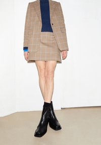 Kostičkovaný hnědý blazer a sukně s námořnickým rolákem. Černé kožené kotníkové boty se čtvercovými špičkami a texturovanými detaily.