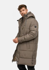 Hnedá púderová bunda s kapucňou, s diagonálnym prešívaním, bočnými vreckami a kontrastnými zipsovými detailmi. Textúra vyzerá hladko a vypchato.