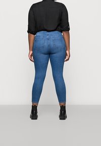 Persona con camisa negra arremangada, jeans skinny azules y botas negras hasta el tobillo, de pie de espaldas sobre un fondo liso.