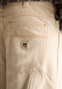 Poche arrière d'un pantalon en denim beige avec des coutures visibles et une petite étiquette de marque Pull&Bear cousue sur la poche.